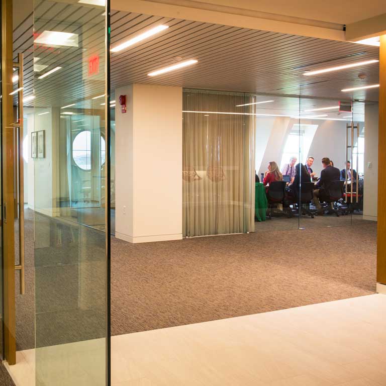 Looking through glass doors at a tan-carpeted open space and a conference room filled with people beyond.