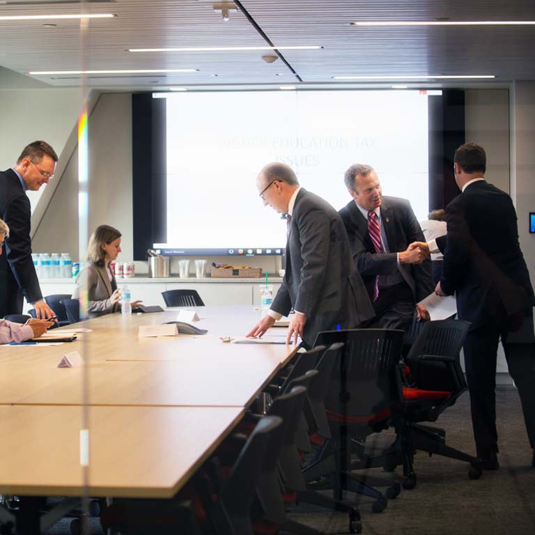 Six men and women in business suits stand talking around a conference table.